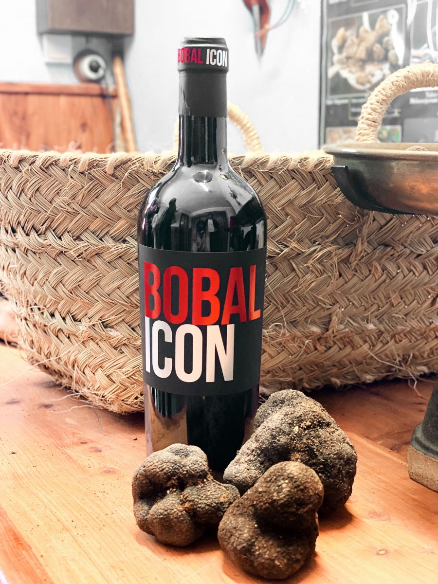 Una botella de vino tinto con etiqueta negra y letras rojas y blancas está colocada sobre una mesa de madera junto a trufas de textura rugosa, con un fondo de cesta tejida en un ambiente ecológico.