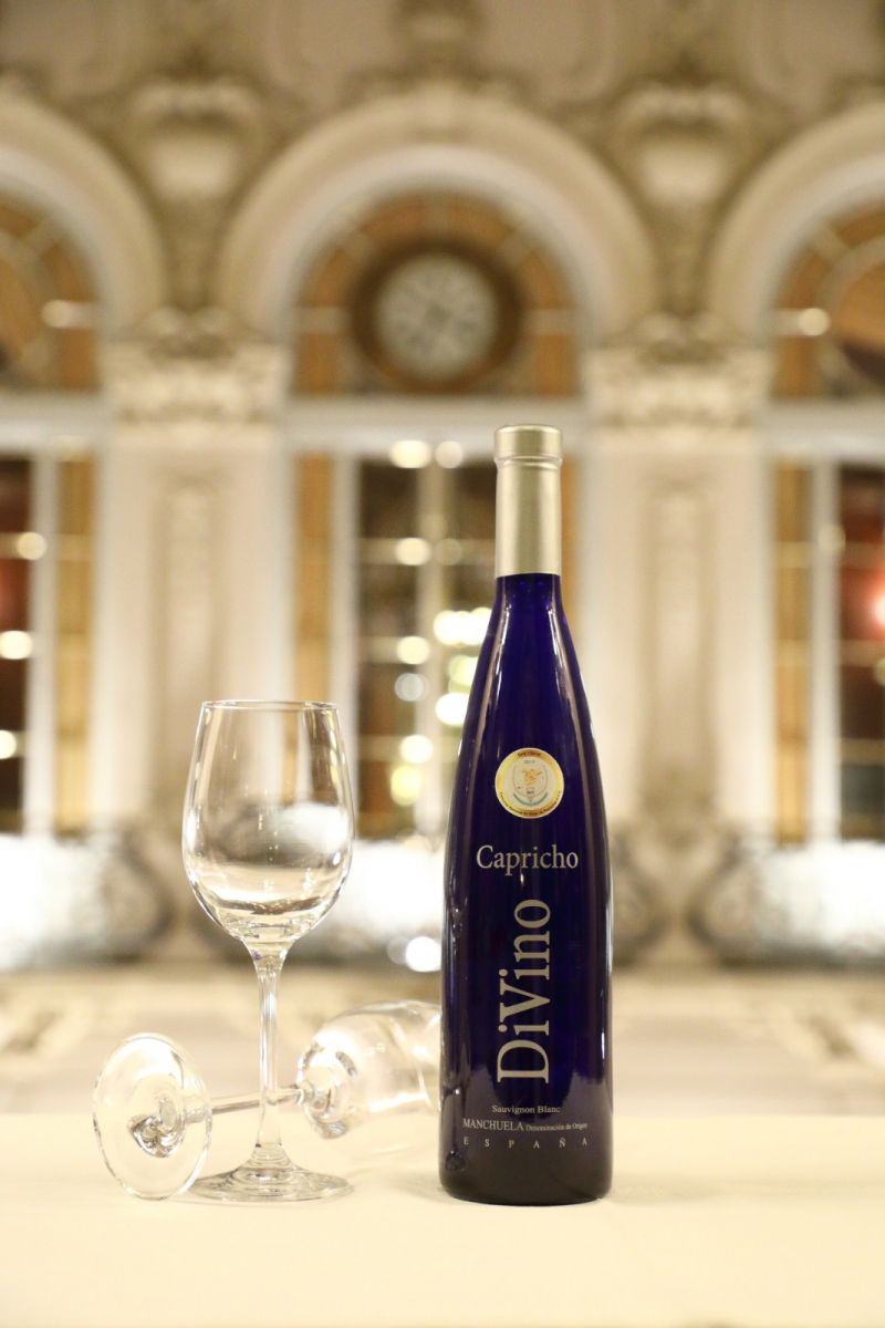 Botella de vino DiVino junto a una copa vacía sobre una mesa con fondo de arquitectura elegante, ideal para comprar online.