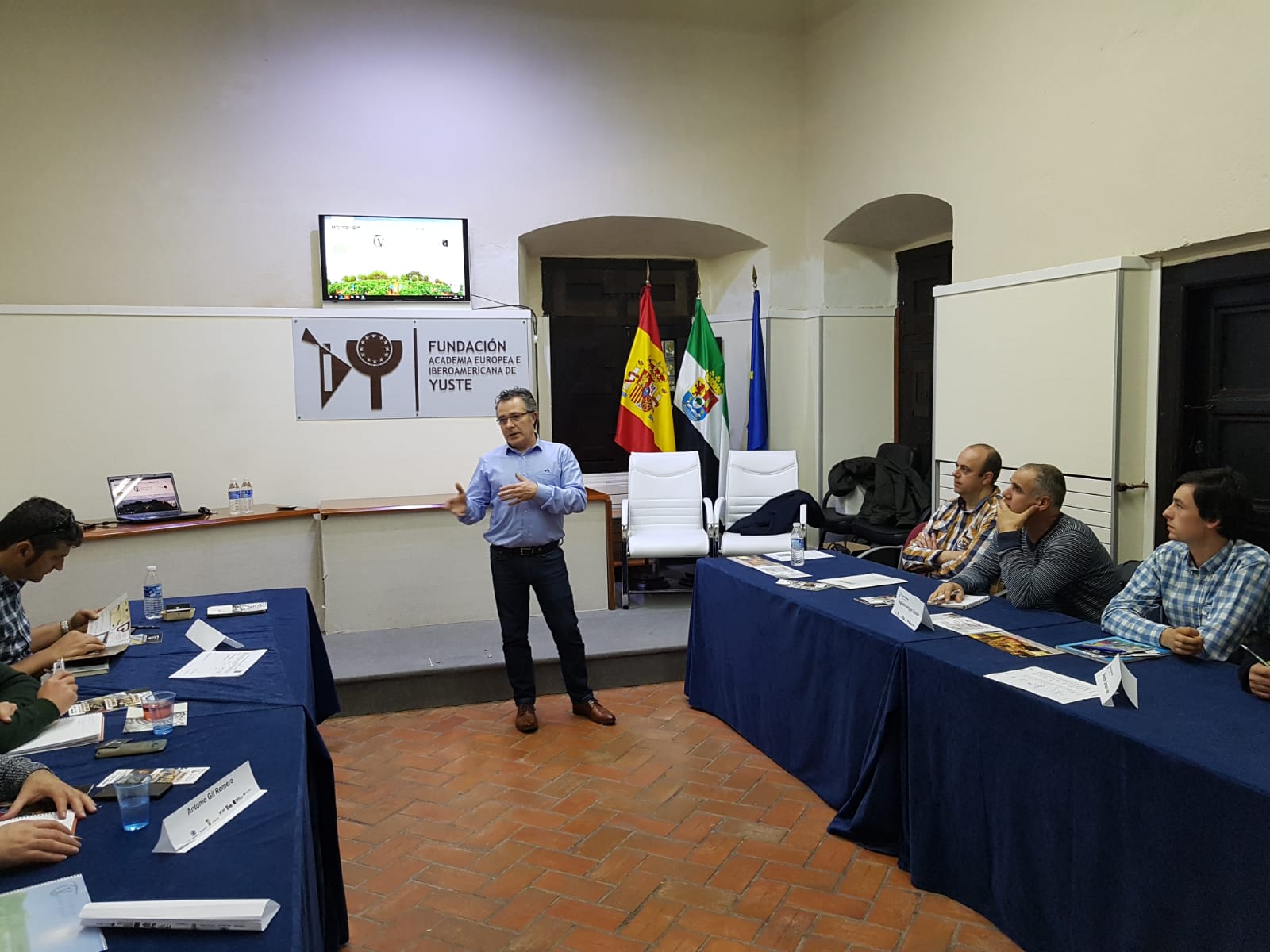 Un hombre de pie da una presentación en una sala con un cartel de la Fundación Academia Europea e Iberoamericana de Yuste, mientras varios hombres sentados lo escuchan, y al fondo se observan banderas, destacándose la participación de la DEO manchuela.