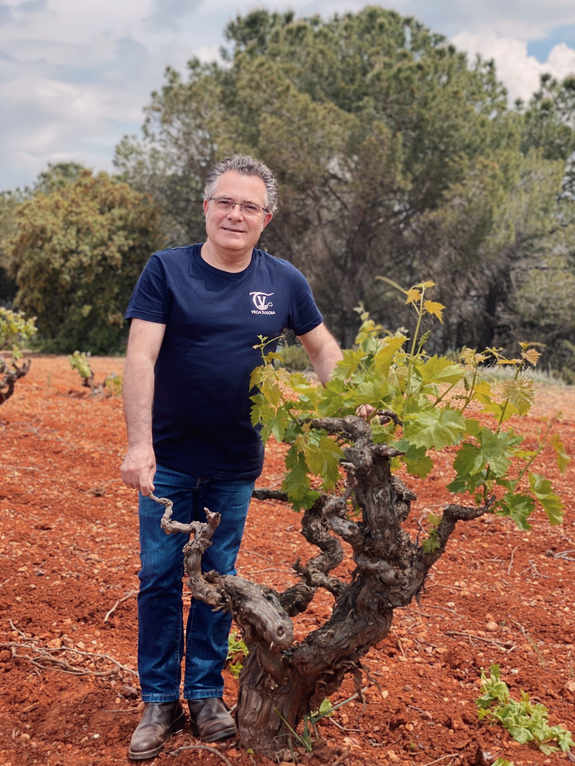 Un hombre sonriente con camiseta azul está junto a una vid en un viñedo de tierra rojiza con un entorno natural y ecológico de fondo.