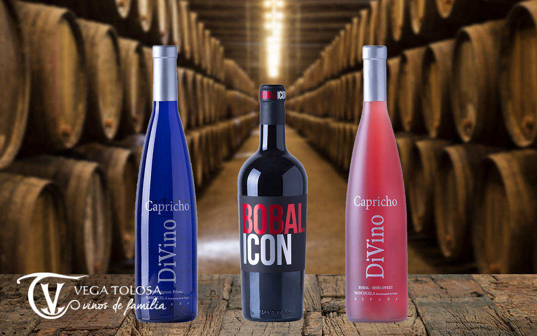 Tres botellas de vino, una azul, una negra y una roja, se presentan frente a una bodega de barricas de madera, destacando el DEO manchuela, reflejando la tradición vinícola de la región.