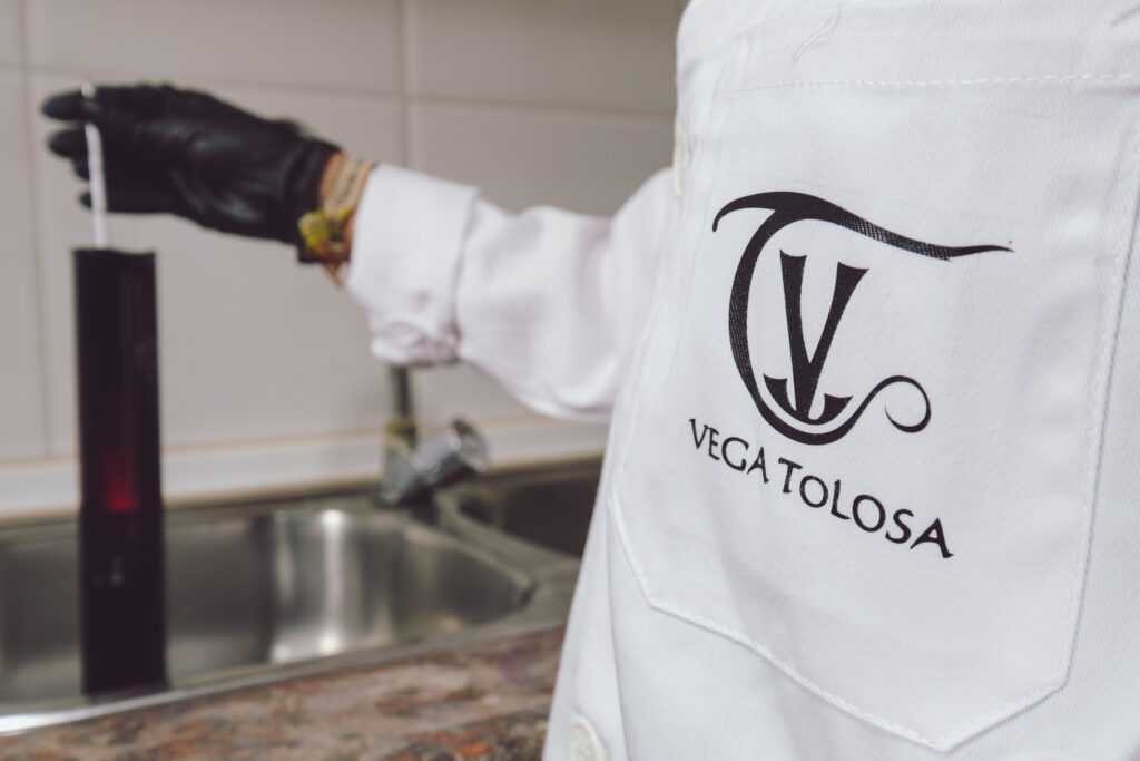Una persona con guante negro sostiene un instrumento de medición en una cocina, mientras porta un uniforme con el logo de una empresa centrada en prácticas ecológicas de la región de La Manchuela.