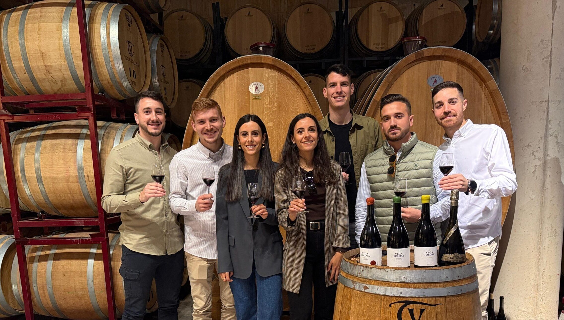 Un grupo de personas sonrientes sostiene copas de vino mientras posa frente a barriles en una bodega, resaltando un ambiente ecológico.