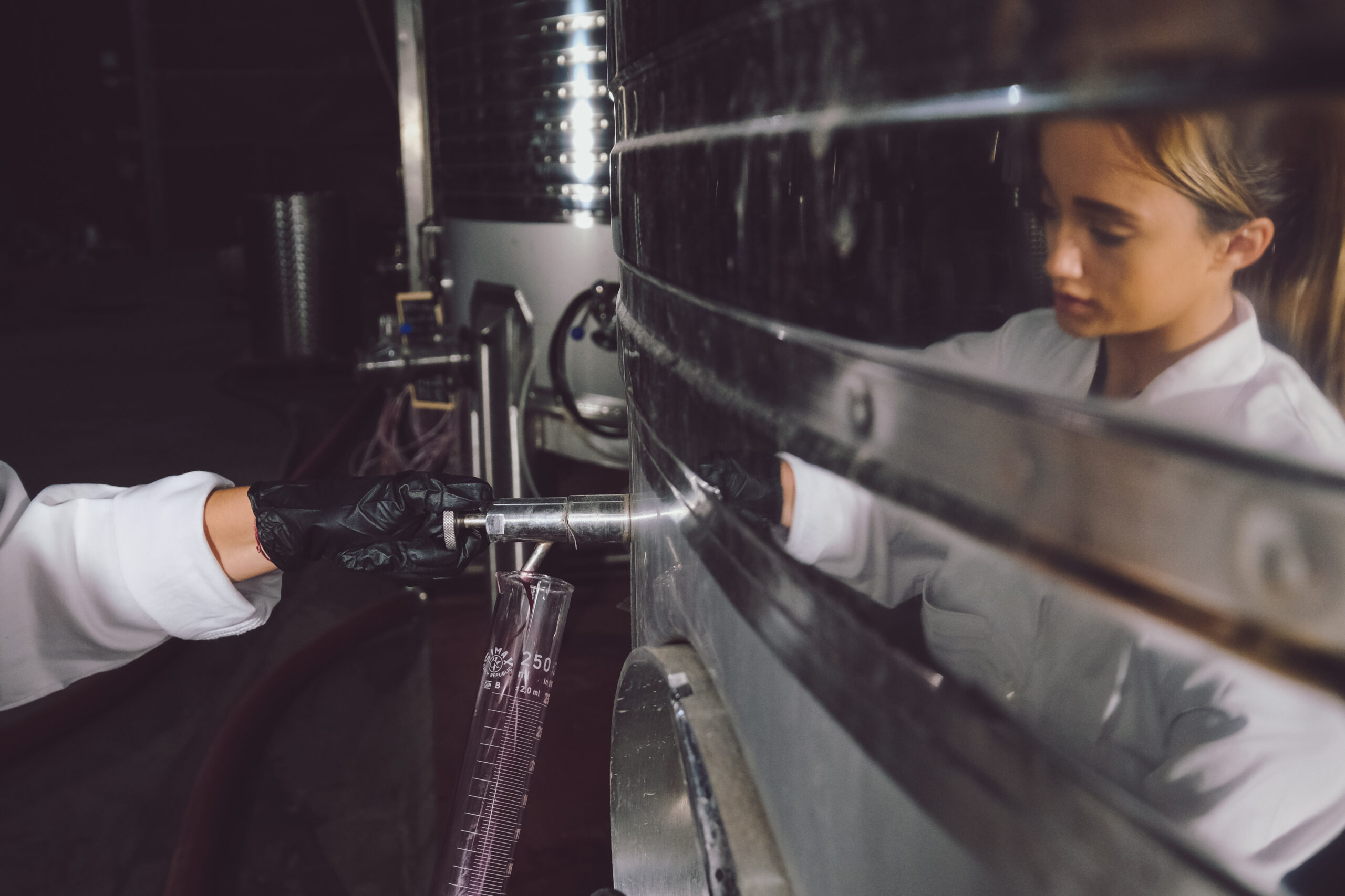 Una mujer con bata blanca y guantes negros extrae líquido de un tanque metálico en un laboratorio, reflejando procesos de calidad y certificados en la producción de manchuela.